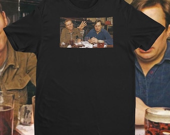 Op de afbeelding: Zwart t-shirt met een afbeelding van twee mannen aan een tafel met drankjes. Eén man zwaait, de ander kijkt vooruit. De afbeelding is een still uit een film of televisieshow.