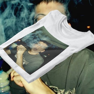 May include: White t-shirt featuring a printed image of a person blowing smoke from a glass pipe.