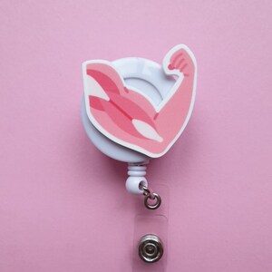 Puede incluir: Portagafete retráctil blanco con un gráfico de brazo muscular rosa. El portagafete tiene una correa de plástico transparente y un clip de metal plateado.
