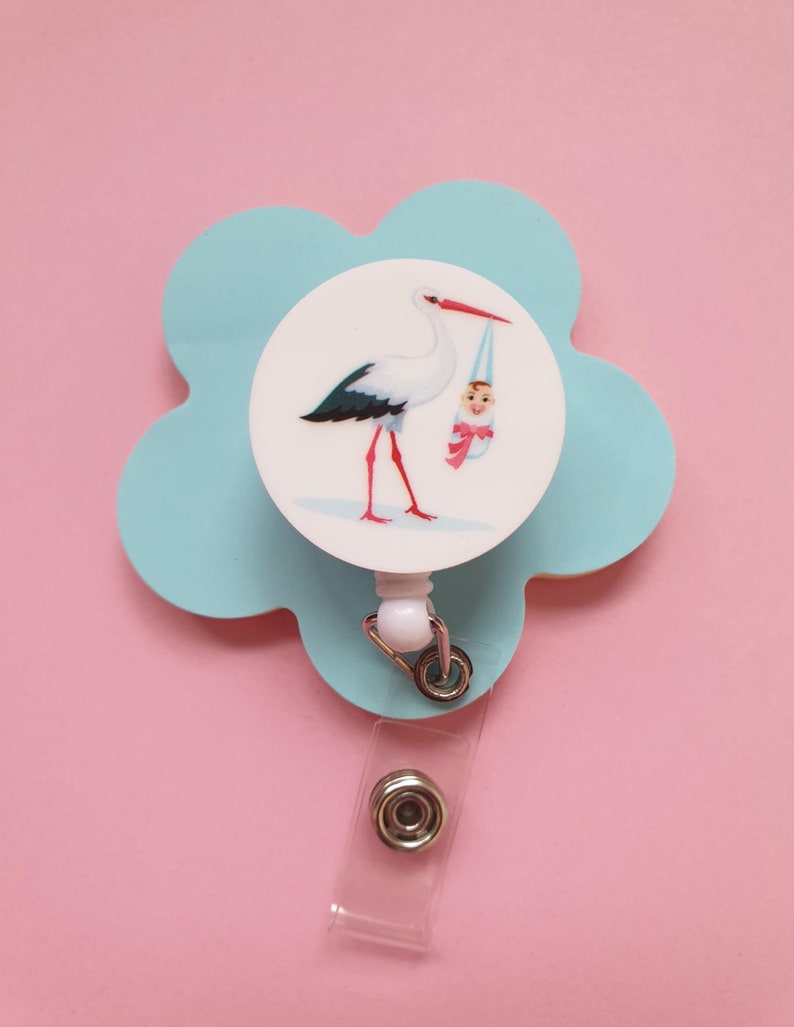 May include: A blue flower-shaped retractable badge holder with a white circle featuring a stork carrying a baby. The badge holder has a clear plastic strap.