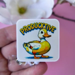 May include: A white square badge with a cartoon duck illustration. The duck is yellow, orange, and white, with the word "PRODUCTIVE" in blue and yellow text above it. The badge is on a retractable reel.