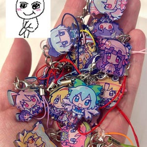 Puede incluir: Una colección de colgantes de personajes de estilo anime con diseños variados, incluyendo personajes con cabello azul, rosa y amarillo. Cada colgante tiene un cierre metálico y un cordón de color.