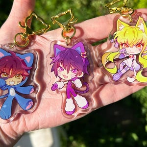 Puede incluir: Tres llaveros de estilo anime con personajes con temática de gatos. Los llaveros tienen cierres dorados. Los personajes visten trajes azules, morados y amarillos con orejas y colas a juego. El fondo es verde borroso.
