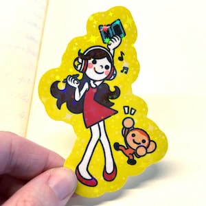May include: A yellow sticker featuring a cartoon girl wearing headphones and a red dress, holding a music player. Musical notes and a small monkey are also on the sticker. The sticker has a holographic, glittery effect.
