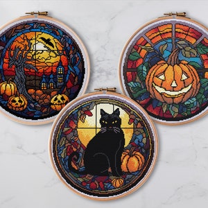 May include: Three round embroidery hoops with colourful stained glass designs. The first hoop features a spooky tree with a crescent moon and pumpkins. The second hoop features a jack-o'-lantern with a toothy grin. The third hoop features a black cat sitting next to a pumpkin.