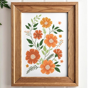 Op de afbeelding: Een ingelijst borduurwerk met kruissteek, met een bloemenmotief met oranje en groene bloemen. De bloemen zijn in een boeketvormig patroon op een witte achtergrond gerangschikt.