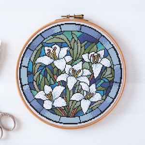 May include: A round embroidery hoop with a cross-stitch design of white lilies in a stained glass style. The lilies are surrounded by blue, green, and brown geometric shapes.