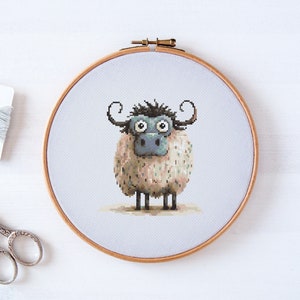 Peut inclure: Broderie au point de croix fantaisiste d'un mouton aux grands yeux et aux cornes bouclées, dans un cercle en bois. Le mouton a un corps duveteux et moucheté et un visage bleu-gris. Fil à broder et ciseaux visibles.