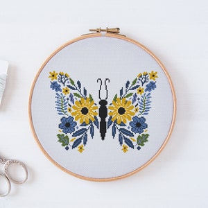 May include: A cross-stitch embroidery hoop featuring a floral butterfly design. The butterfly is crafted from yellow, blue, and green flowers with a black outline. The hoop is made of wood and has a white background.