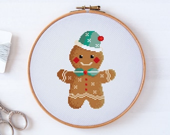 May include: A cross-stitch pattern of a gingerbread man wearing a green and white hat with a red pom-pom. The gingerbread man is smiling and has a green bow tie.