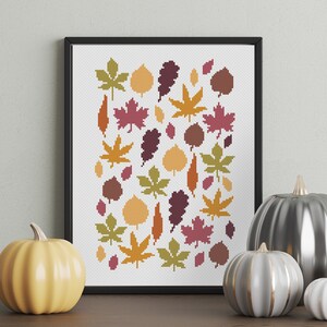 May include: A framed cross-stitch artwork featuring a pattern of colorful autumn leaves in shades of orange, yellow, green, and burgundy. Decorative pumpkins in various sizes and colors are arranged in front of the artwork.