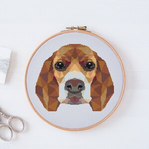 May include: A cross-stitch pattern of a beagle dog's face in a geometric style. The dog's fur is brown, white, and black. The dog's eyes are black and the nose is brown. The dog's mouth is closed and the tongue is not visible.