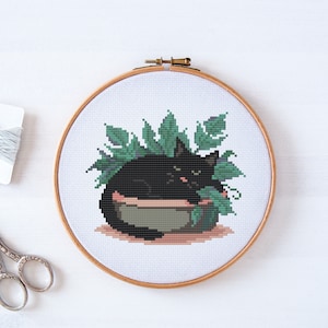 May include: Cross stitch pattern of a black cat curled up in a pot of green plants. The cat has a sleepy expression.