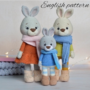 May include: Three crocheted bunny plushies in different sizes, wearing colorful scarves and outfits. The largest bunny is wearing an orange dress, the middle bunny is wearing a blue sweater, and the smallest bunny is wearing a yellow sweater.  The bunnies are all standing on a white surface.  English pattern.