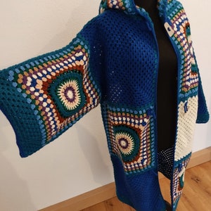 Könnte beinhalten: Ein langer, blauer Häkelcardigan mit Kapuze. Der Cardigan hat ein weißes und mehrfarbiges Häkeldesign an den Ärmeln und vorne.