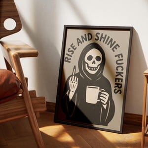 May include: A framed poster with a black border features a Grim Reaper illustration holding a mug and giving the middle finger. The text "RISE AND SHINE FUCKERS" is printed around the image in a bold, black font. The poster is leaning against a wall.