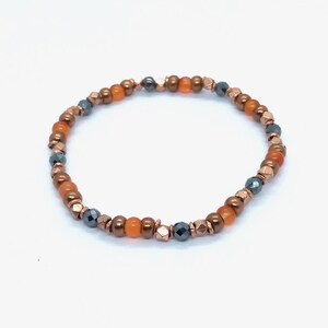Peut inclure: Un bracelet de perles composé d'un mélange de perles orange, cuivrées et gris foncé. Le bracelet est composé de perles rondes et facettées, enfilées sur un cordon élastique. Il est conçu comme un accessoire de mode.