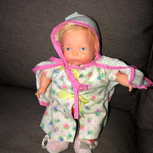 May include: A vintage baby doll with blonde hair and blue eyes, wearing a floral print outfit with pink trim and a matching bonnet. The doll's outfit includes a cape and matching booties. The doll is posed on a dark gray surface.