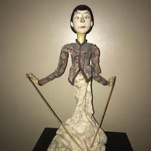 May include: A vintage Asian puppet with a white face, black hair, and a patterned fabric outfit. The puppet is holding two wooden sticks in its hands.