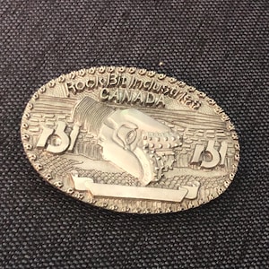 May include: Silver-coloured oval belt buckle featuring the text "Rock Bit Industries CANADA" and the letters "B.I." flanking a detailed machine illustration. The buckle has a textured border.