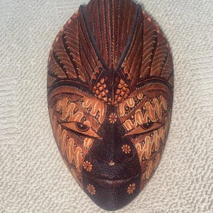May include: An oval-shaped wooden mask featuring intricate hand-painted patterns in shades of dark brown, tan, and orange. The design includes stylised floral motifs, fine dot work, and a prominent central crest at the top, with almond-shaped eyes.
