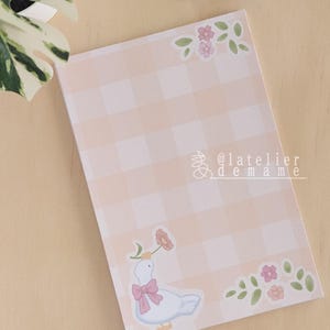 Può includere: Un blocco note rettangolare con un motivo a quadretti vichy color pesca chiaro e bianco. Il design include piccoli gruppi di fiori rosa e verdi negli angoli e un'oca bianca con un fiocco rosa che tiene un fiore. Il testo @latelierdemame è visibile.
