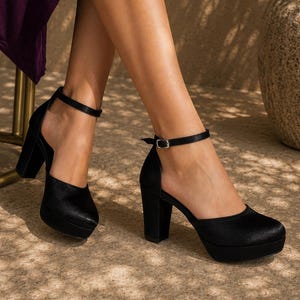 May include: Black platform heels with ankle straps. The shoes have a rounded toe and a thick block heel. The platform sole adds height. The shoes are made of a soft, textured material. The ankle straps have a small silver buckle.