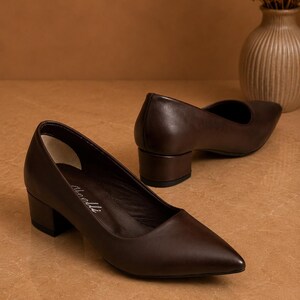 Peut inclure: Une paire d'escarpins en cuir marron fonc&eacute; avec un bout pointu et un talon bloc. Les chaussures sont pr&eacute;sent&eacute;es sur une surface marron clair. La hauteur du talon est d'environ 5 cm.