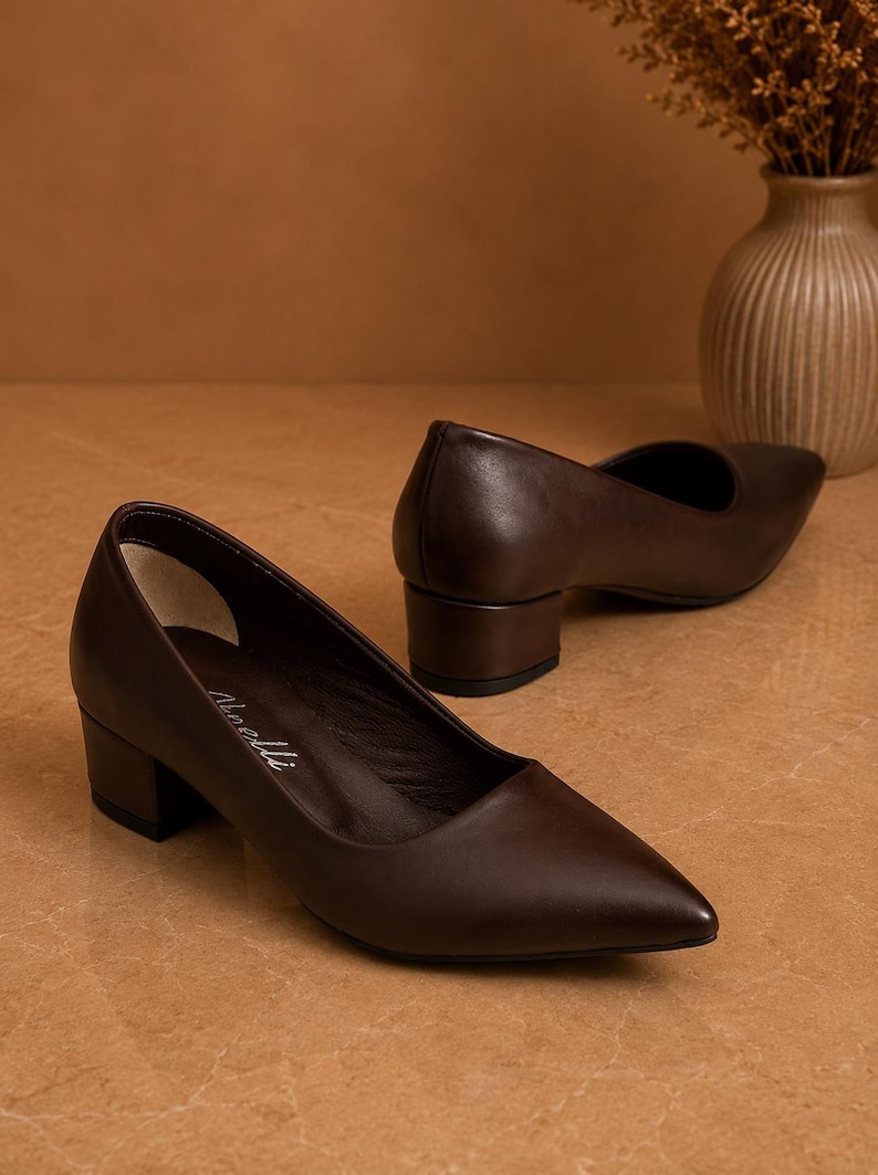Peut inclure: Une paire d'escarpins en cuir marron fonc&eacute; avec un bout pointu et un talon bloc. Les chaussures sont pr&eacute;sent&eacute;es sur une surface marron clair. La hauteur du talon est d'environ 5 cm.