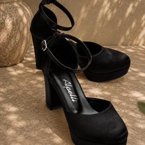 May include: Black platform heels with ankle straps. The shoes have a rounded toe and a thick block heel. The brand name "Apelli" is visible on the insole. The shoes are made of a soft, velvety material.