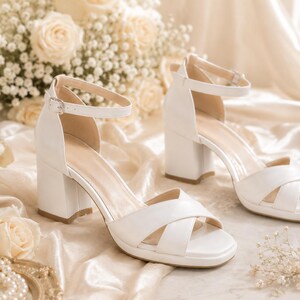 May include: A pair of white, block-heeled sandals with ankle straps. The sandals have a criss-cross design over the toes and are set against a backdrop of cream-colored roses and fabric. The heels appear to be approximately 3 inches high.