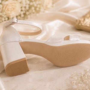 May include: Ivory-colored sandal with a block heel and ankle strap. The shoe features a platform sole and a textured, light brown outsole. The heel measures approximately 3 inches.