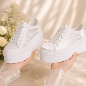 Può includere: Un paio di sneakers platform bianche con chiusura a lacci e un rivestimento in pizzo floreale testurizzato sulla tomaia. Le scarpe presentano una spessa suola in gomma bianca con un motivo a coste orizzontali lungo il lato della base.