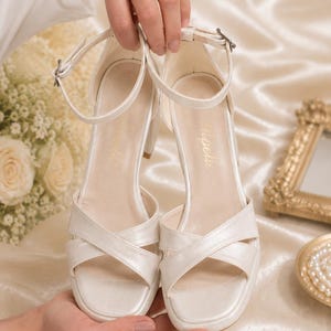May include: Ivory-colored bridal sandals with a platform sole and crisscross straps. The heels are a medium height. The sandals have an ankle strap with a buckle closure. The word "Tipelli" is printed on the insole.