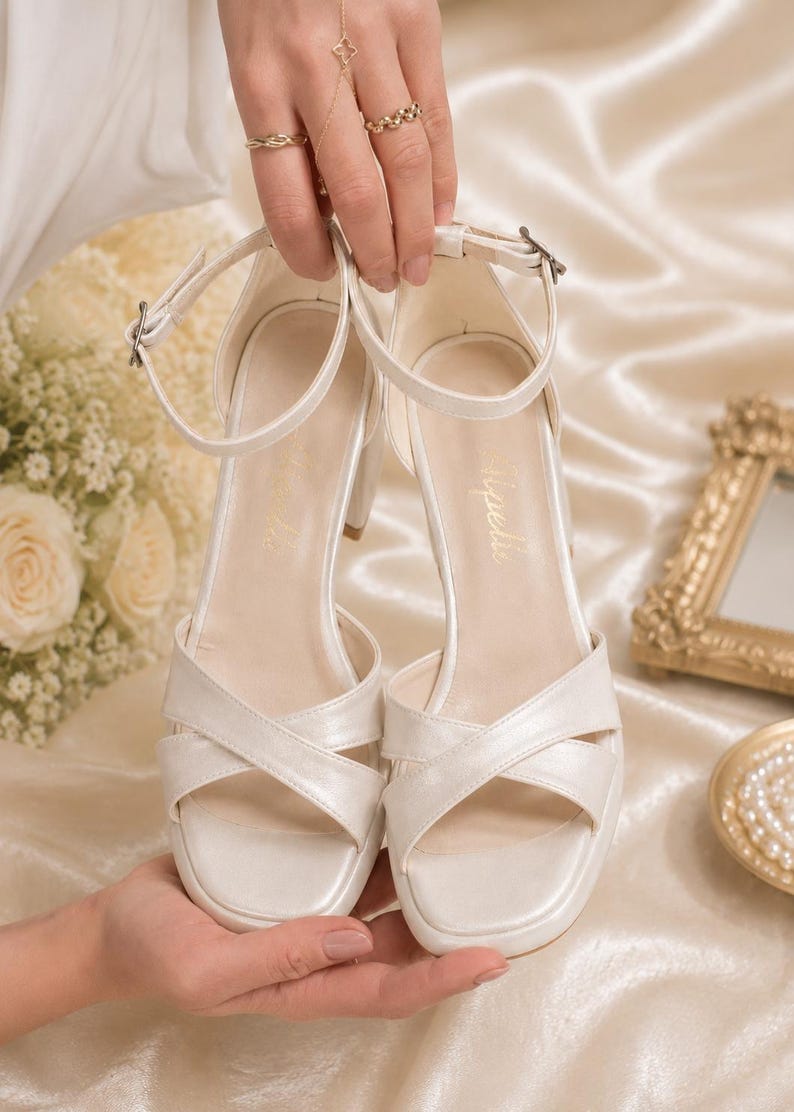 May include: Ivory-colored bridal sandals with a platform sole and crisscross straps. The heels are a medium height. The sandals have an ankle strap with a buckle closure. The word "Tipelli" is printed on the insole.