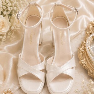 May include: Ivory-colored heeled sandals with a crisscross strap design and ankle straps. The block heels and square toes offer a modern look. The sandals are displayed on a satin surface, with floral and pearl accents.