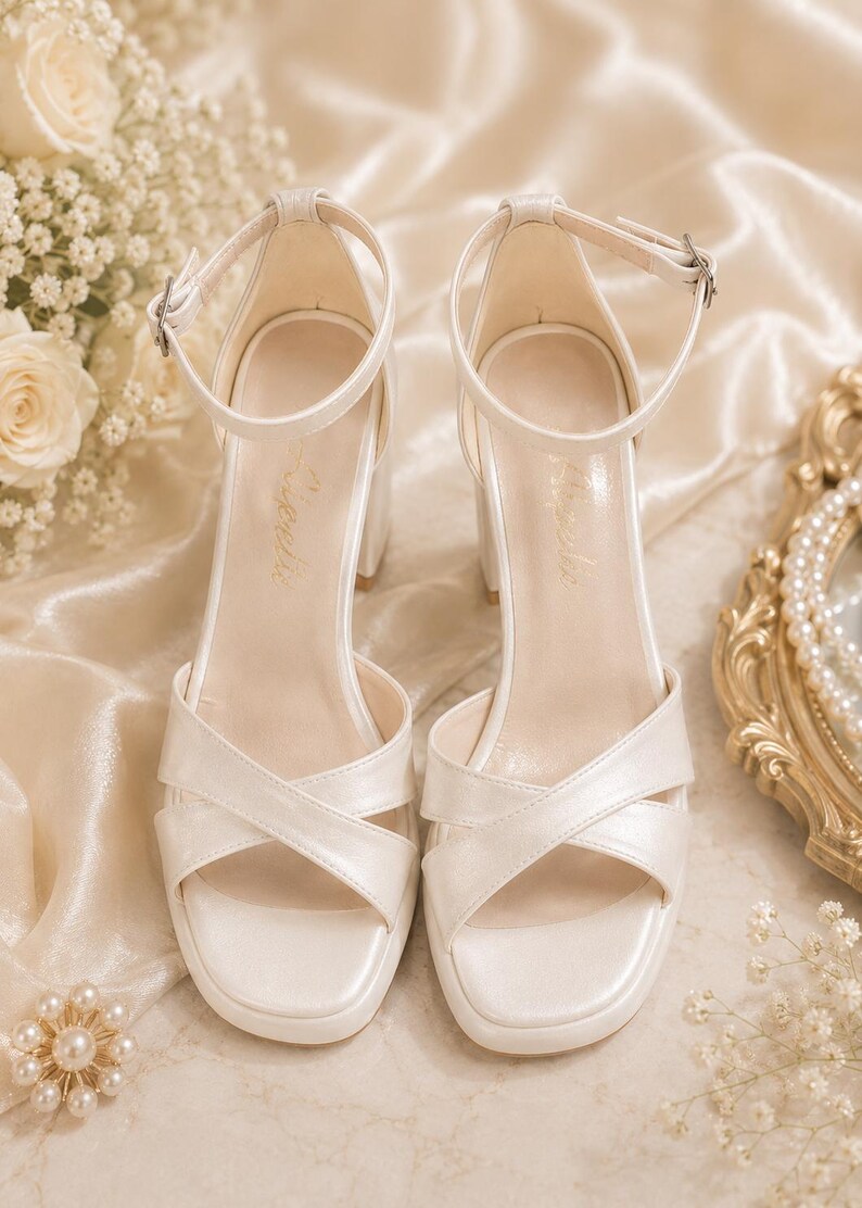 May include: Ivory-colored heeled sandals with a crisscross strap design and ankle straps. The block heels and square toes offer a modern look. The sandals are displayed on a satin surface, with floral and pearl accents.
