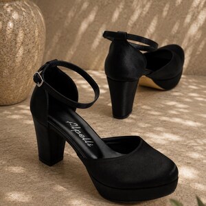 May include: Black platform heels with ankle straps. The shoes have a rounded toe and a thick, block heel. The heels are approximately 10 cm high. The shoes are made of a smooth, satin-like material.
