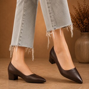 Peut inclure: Escarpins en cuir marron fonc&eacute; &agrave; bout pointu et petit talon bloc. Les chaussures sont port&eacute;es avec un jean en denim d&eacute;lav&eacute; clair avec des ourlets effiloch&eacute;s. L'arri&egrave;re-plan pr&eacute;sente une palette de couleurs neutres avec un vase de fleurs s&eacute;ch&eacute;es.