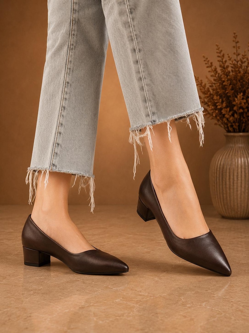 Peut inclure: Escarpins en cuir marron fonc&eacute; &agrave; bout pointu et petit talon bloc. Les chaussures sont port&eacute;es avec un jean en denim d&eacute;lav&eacute; clair avec des ourlets effiloch&eacute;s. L'arri&egrave;re-plan pr&eacute;sente une palette de couleurs neutres avec un vase de fleurs s&eacute;ch&eacute;es.