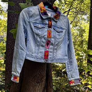 May include: A light blue denim jacket featuring a collar and cuffs accented with multi-colored patterned fabric patches. The front button placket is decorated with a vertical row of small, rectangular fabric patches, each displaying a stitched heart design.