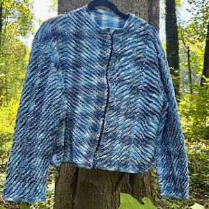 May include: A long-sleeve jacket featuring a textured, chevron-patterned surface in varying shades of blue and white. The garment has a round neckline, a straight hem, and a dark vertical zipper closure running down the center front of the piece.