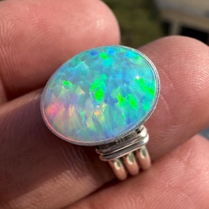 May include: A silver ring featuring an oval opal. The opal exhibits a striking iridescence, with hues of blue, green, and pink. The ring's design is minimalist, highlighting the gemstone's natural colours and patterns.
