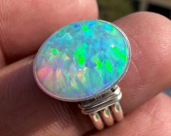 May include: Oval-shaped opal ring with a silver band. The opal displays a vibrant play of color, with flashes of blue, green, and pink. The ring's setting is a simple, yet elegant design, showcasing the gemstone's natural beauty.
