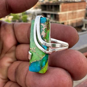 May include: A rectangular ring featuring a vibrant green and turquoise stone with copper-colored veining. The stone is set in a polished silver metal frame that wraps around the top and sides, secured by a split silver band that encircles the finger.