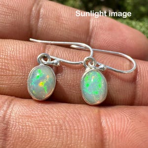 May include: A pair of silver drop earrings featuring oval-shaped opals. The opals display a play of color with green, yellow, and orange hues. The earrings are set in a simple silver setting with a hook closure.
