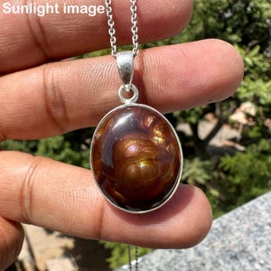 May include: A silver pendant necklace featuring a large, oval-shaped, polished gemstone with brown and gold tones. The gemstone is set in a silver bezel and hangs from a delicate silver chain. The pendant is held up by a person's fingers.