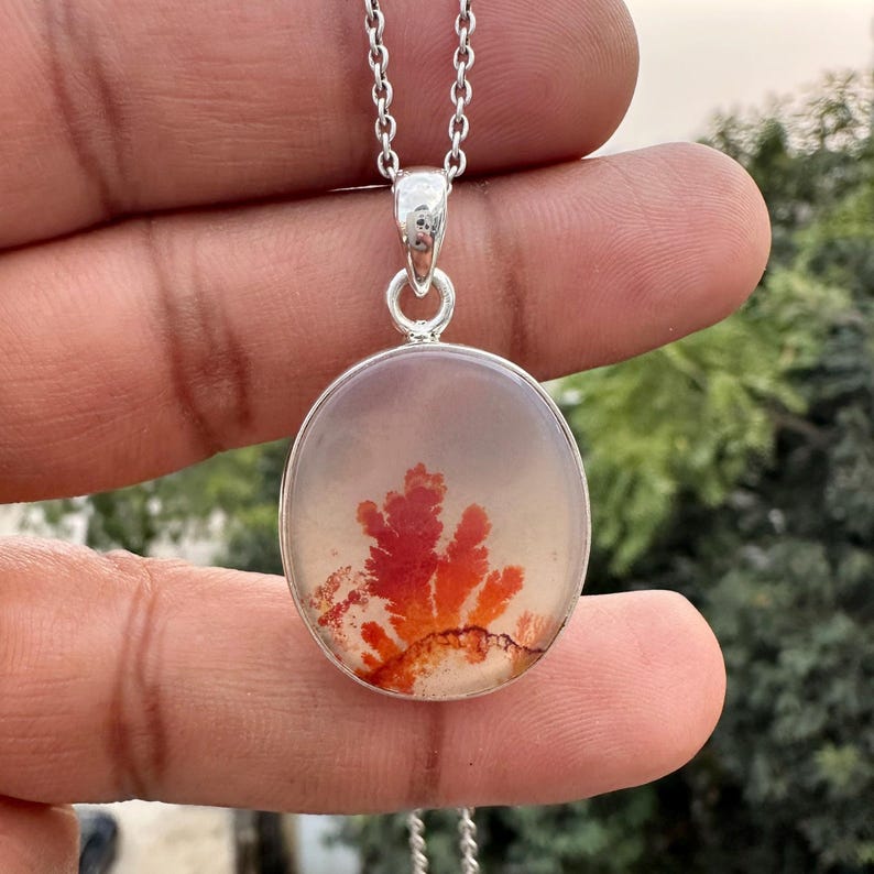 May include: A silver pendant necklace featuring a round, clear stone with a vibrant red and orange dendritic pattern. The pendant hangs from a delicate silver chain, showcasing the natural beauty of the stone's unique design. The necklace is a piece of jewelry.