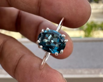 May include: A silver ring featuring a large, oval-cut, deep blue gemstone held in place by six prongs. The faceted stone displays a dark, reflective surface, and the band is made of a smooth, polished silver metal, creating a simple and elegant design.