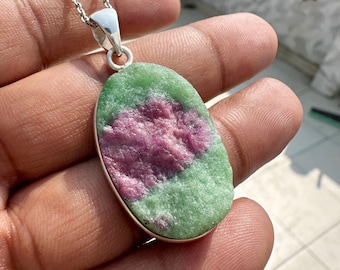 May include: A silver pendant necklace featuring a large, oval-shaped gemstone. The stone has a green and pink color scheme, with a textured surface. The pendant is suspended from a delicate silver chain.